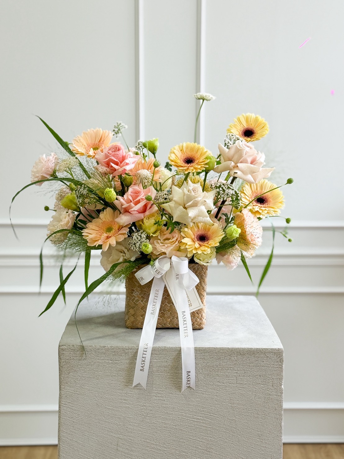 A Sweet 4 Love & Quicksand Roses Pastel Basket, filled with pastel blooms—including roses, gerbera daisies, and greenery—rests on a white plinth. Delicate “NOSTELLE” ribbons adorn the woven basket for an elegant touch.