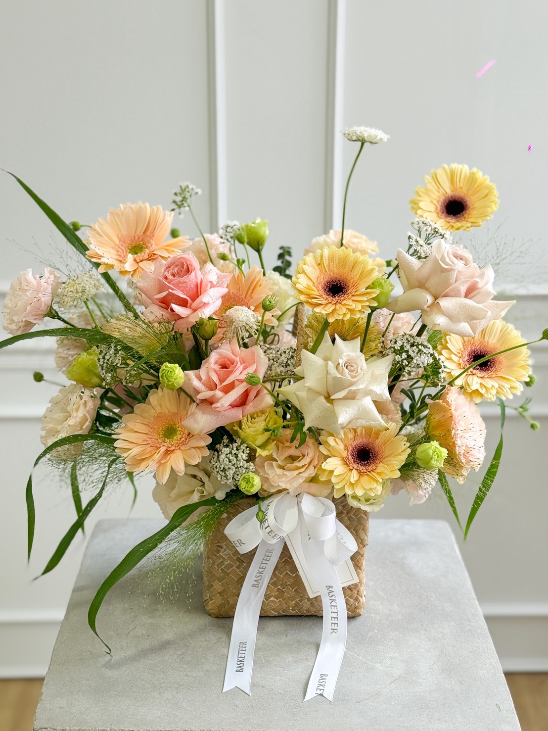 The Sweet 4 Love & Quicksand Roses Pastel Basket features pink roses, yellow gerbera daisies, and white filler flowers with greenery, all arranged in a basket topped with a printed white ribbon. Perfect for adding charm to any space.