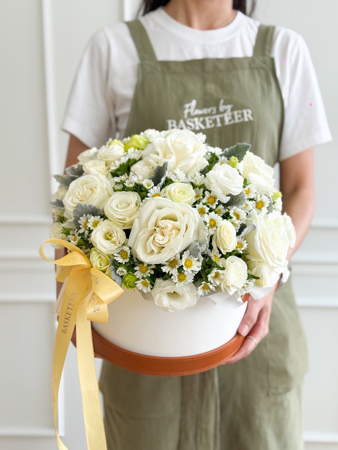 A person in a green apron holds a Playa Blanca Roses & White Daisy Leather Flower Box, filled with white roses, small daisies, and greenery, accented with a yellow ribbon. The apron reads “Flowers by BASKETEER.”.
