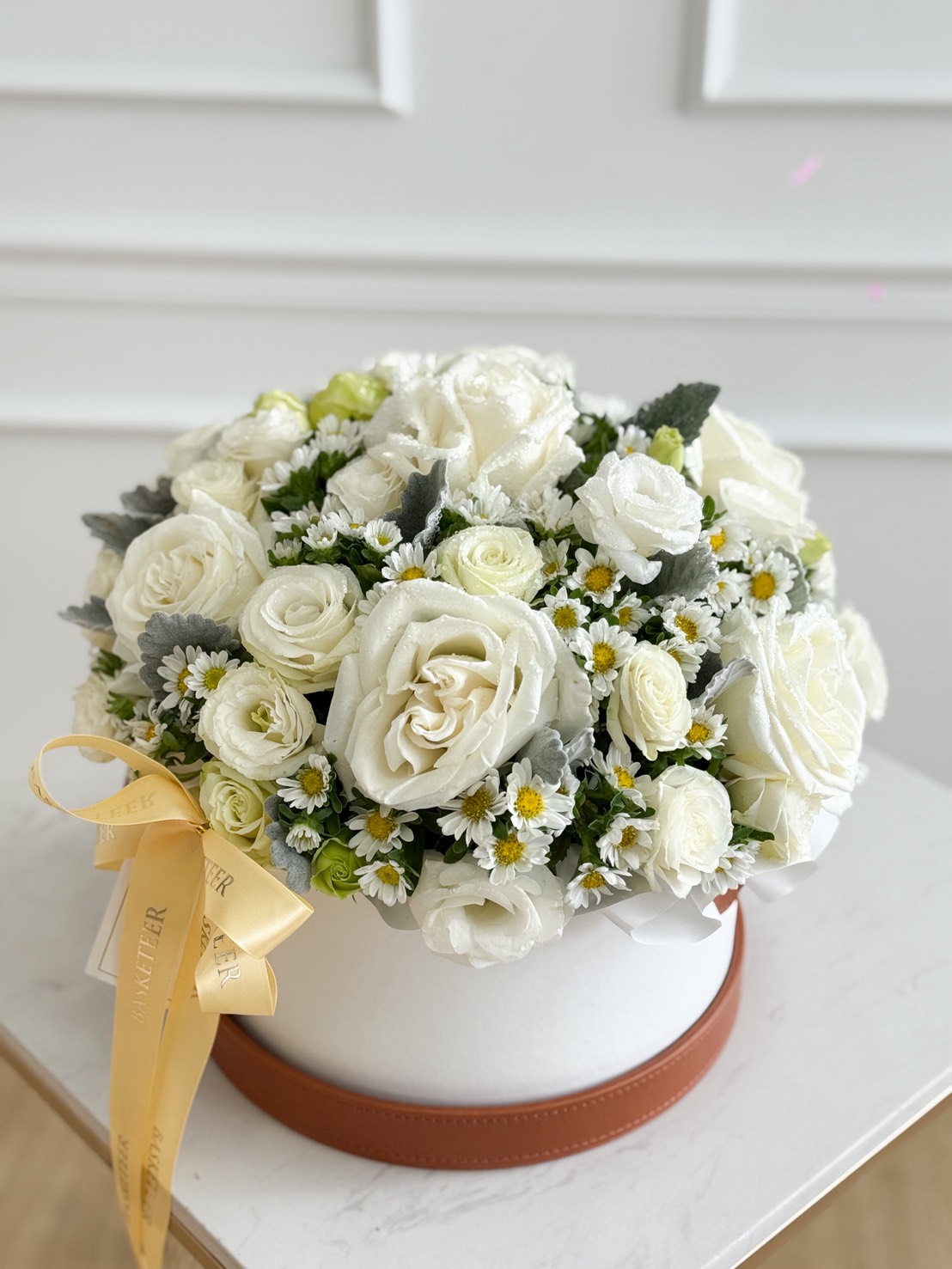 The Playa Blanca Roses & White Daisy Leather Flower Box features elegant white roses, delicate daisies, and lush foliage in a round white box with a yellow ribbon, all beautifully displayed on a marble table against a panelled wall.