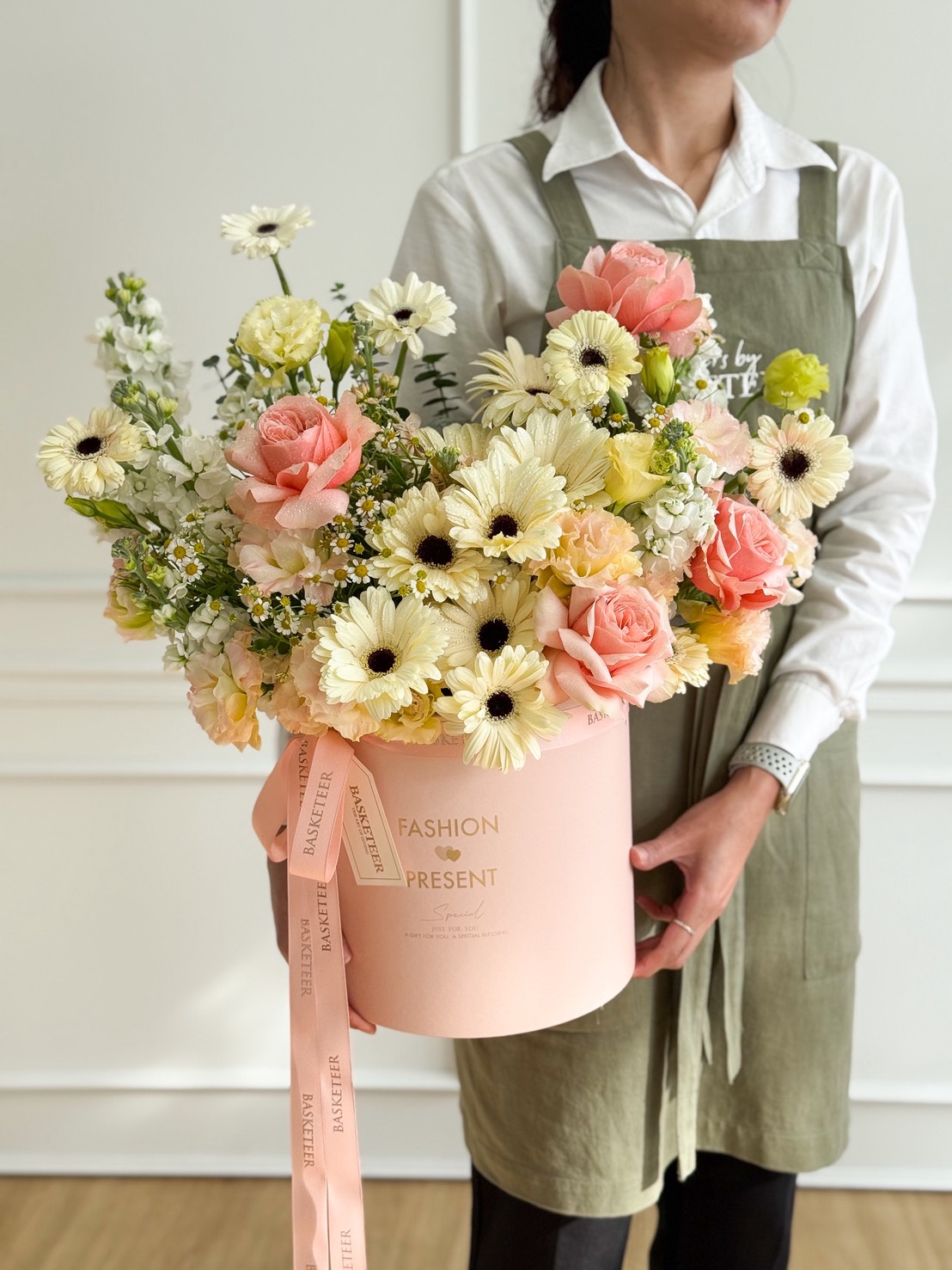 A woman in a green apron holds a large pastel pink hatbox filled with Candlelight Roses & Gerberas in a Pink Velvet Box, cream and pink blooms arranged beautifully, with a ribbon reading 