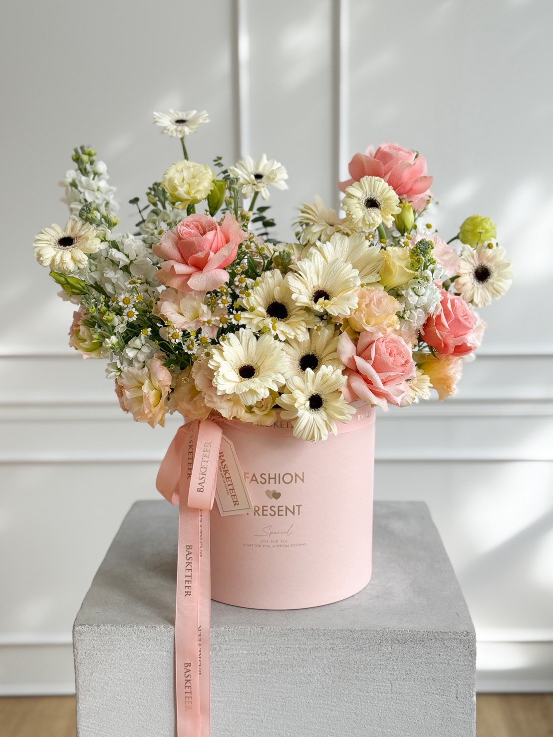 A pink hatbox filled with Candlelight Roses & Gerberas in a Pink Velvet Box, alongside cream and white blooms, sits on a grey pedestal. The box features a pink ribbon and elegant gold writing on the front.
