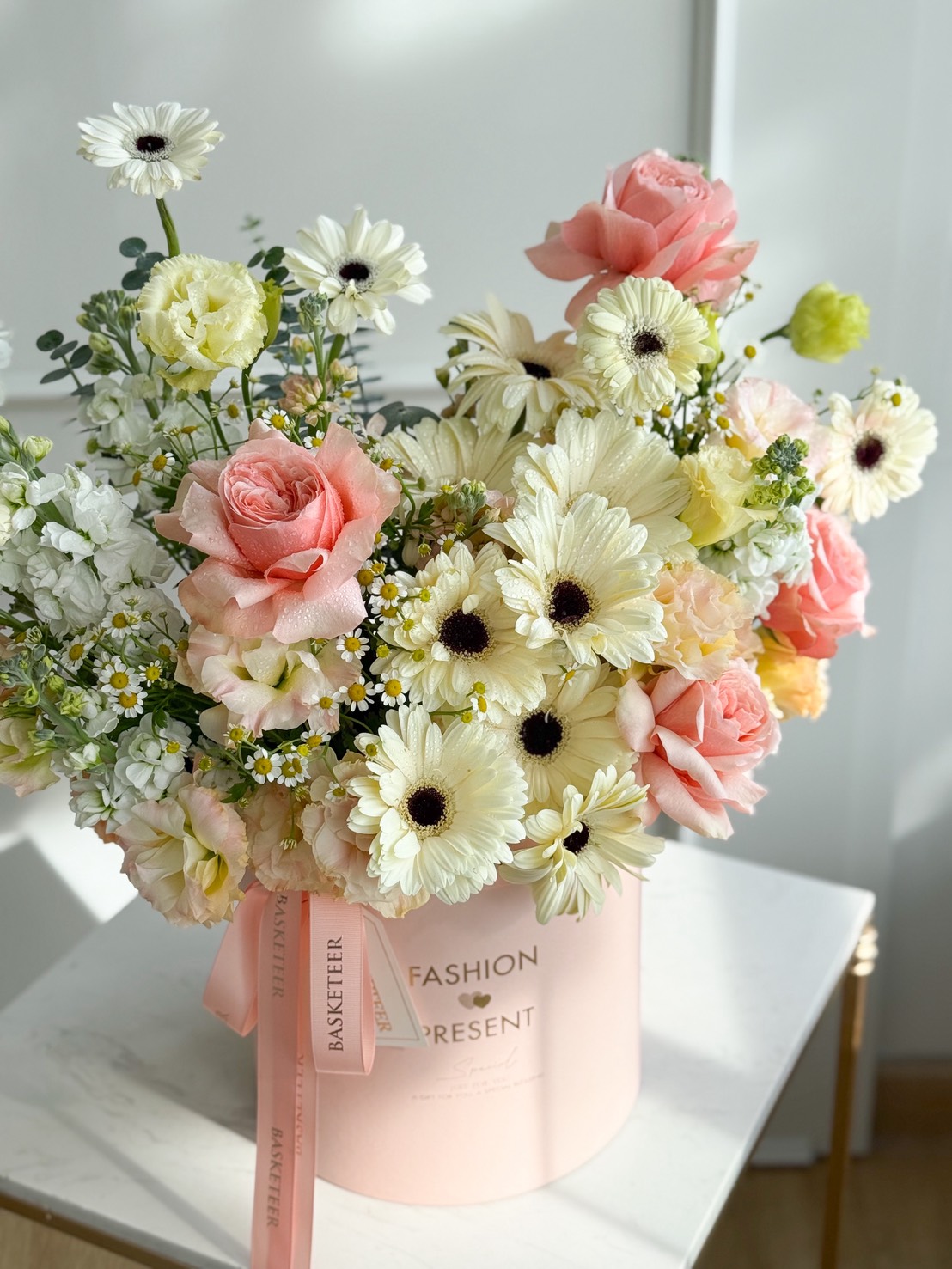 Candlelight Roses & Gerberas in Pink Velvet Box: A pastel flower arrangement with pale yellow gerbera daisies, white ranunculus, and blush pink roses in a round pink box, displayed on a white table in soft natural light.