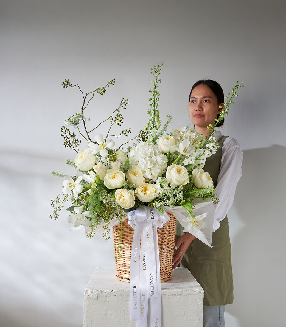 Full view of white and green flower basket with imported Coldplay Roses, hydrangeas, and delphinium tied with white satin ribbon.