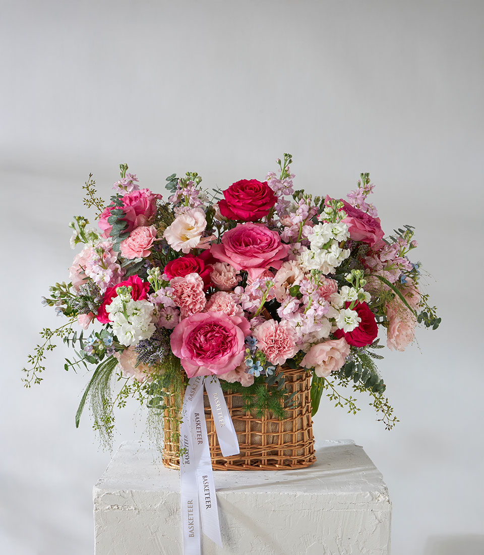 Full view of pink and red flower basket with Nuage and Full Monty Roses, carnations, and white fillers arranged in a woven basket tied with satin ribbon.
