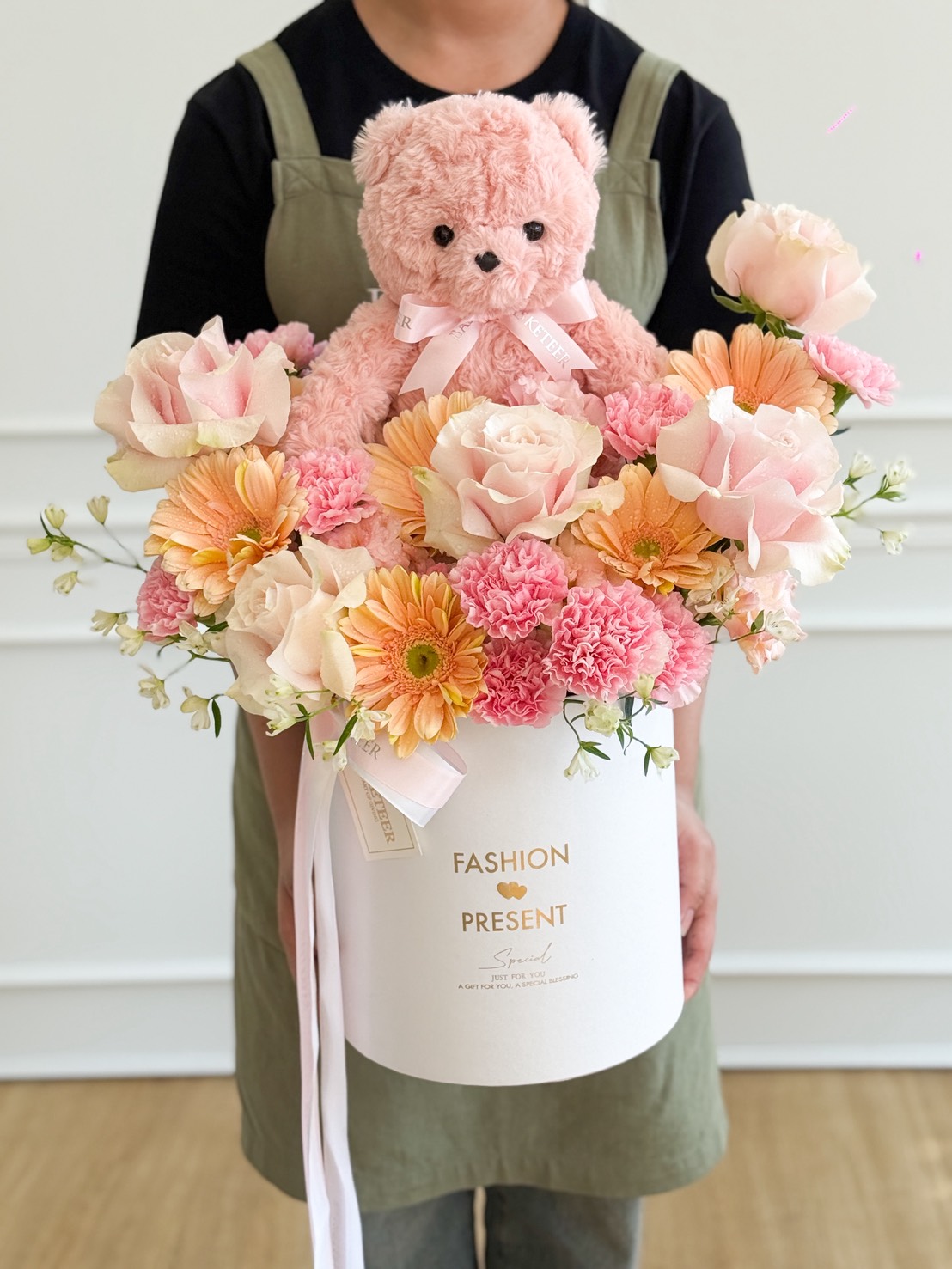 A person in an apron holds a Pink Mondial Roses & Blush Teddy Bear Flower Box, filled with pink and peach blooms and a pink teddy bear. The white box is labelled 