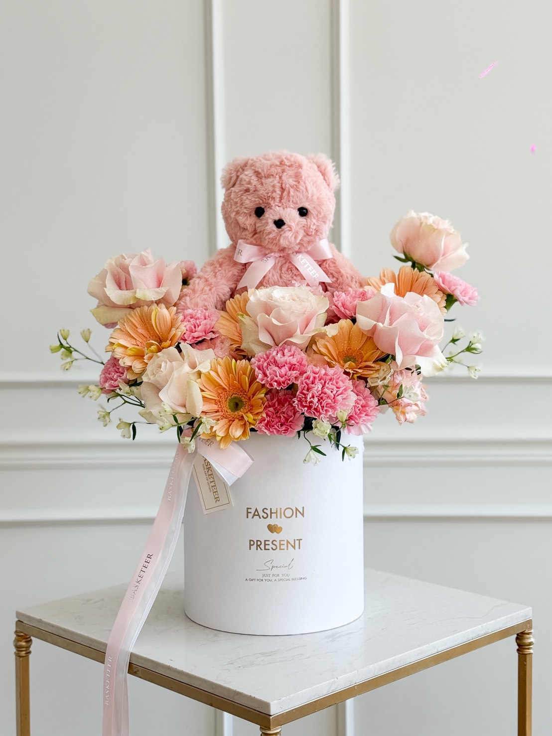 A Blush Teddy Bear Flower Box, featuring a pink teddy bear, sits in the centre of a white hatbox filled with Pink Mondial Roses and carnations, placed on a small table against a white panelled wall.