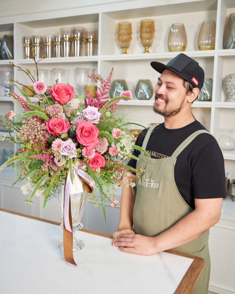 Florist from Flowers by Basketeer smiling beside a pink and lavender flower arrangement