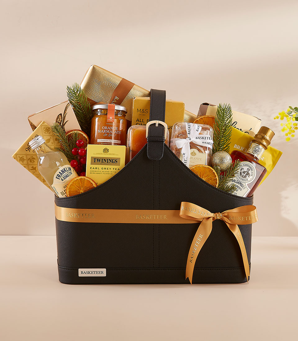 A black gift hamper with a gold ribbon holds tea, honey, biscuits, preserves, dried orange slices, and assorted gourmet treats, decorated with evergreen sprigs and a festive, elegant presentation.
