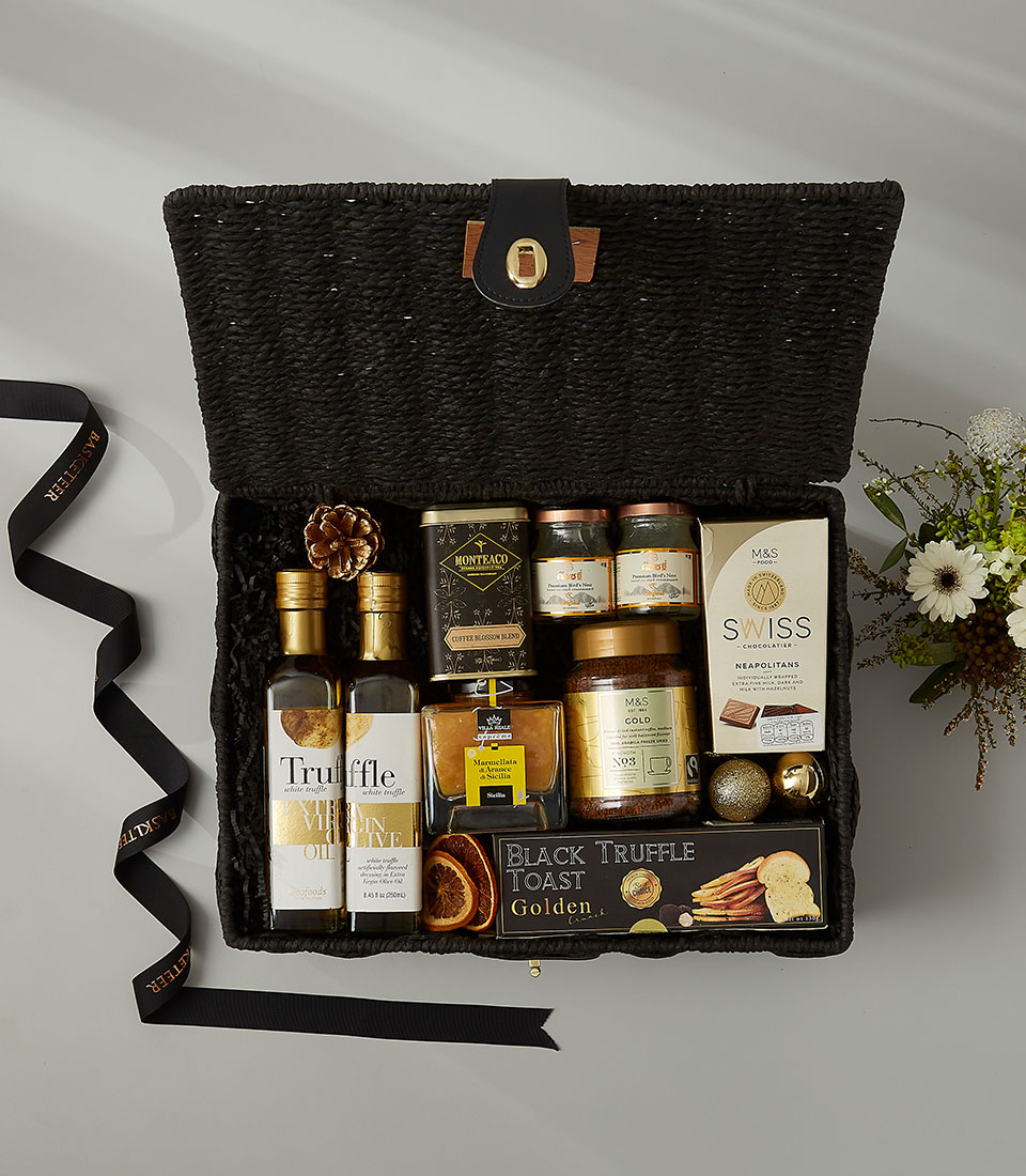 A black woven hamper filled with gourmet items including truffle oils, honey, mustards, jams, Swiss chocolates, biscuits, a tin, and a small festive floral arrangement nearby. A black ribbon is placed beside the hamper.