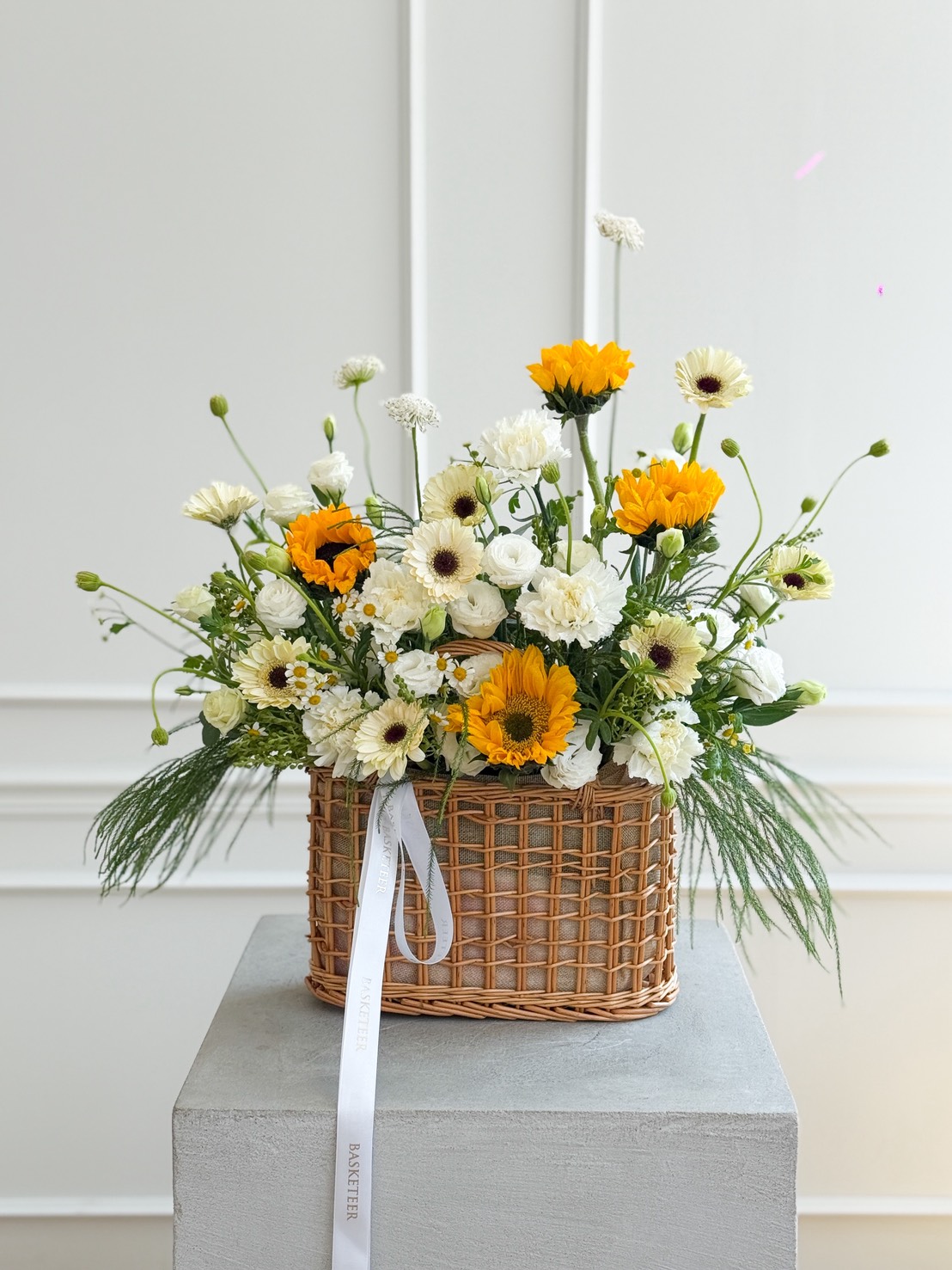 A wicker basket filled with a Sunflowers & White Gerbera Daisy Basket Bouquet and assorted greenery sits on a grey pedestal against a white wall, with a white ribbon tied to the basket’s handle.