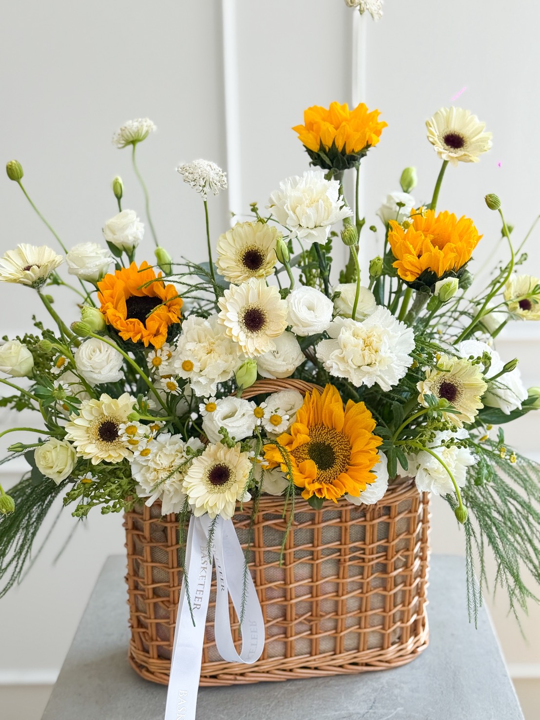 A Sunflowers & White Gerbera Daisy Basket Bouquet arranged with sunflowers, white roses, gerberas, and lush greenery in a wicker basket tied with a white ribbon, set on a grey surface against a light background.