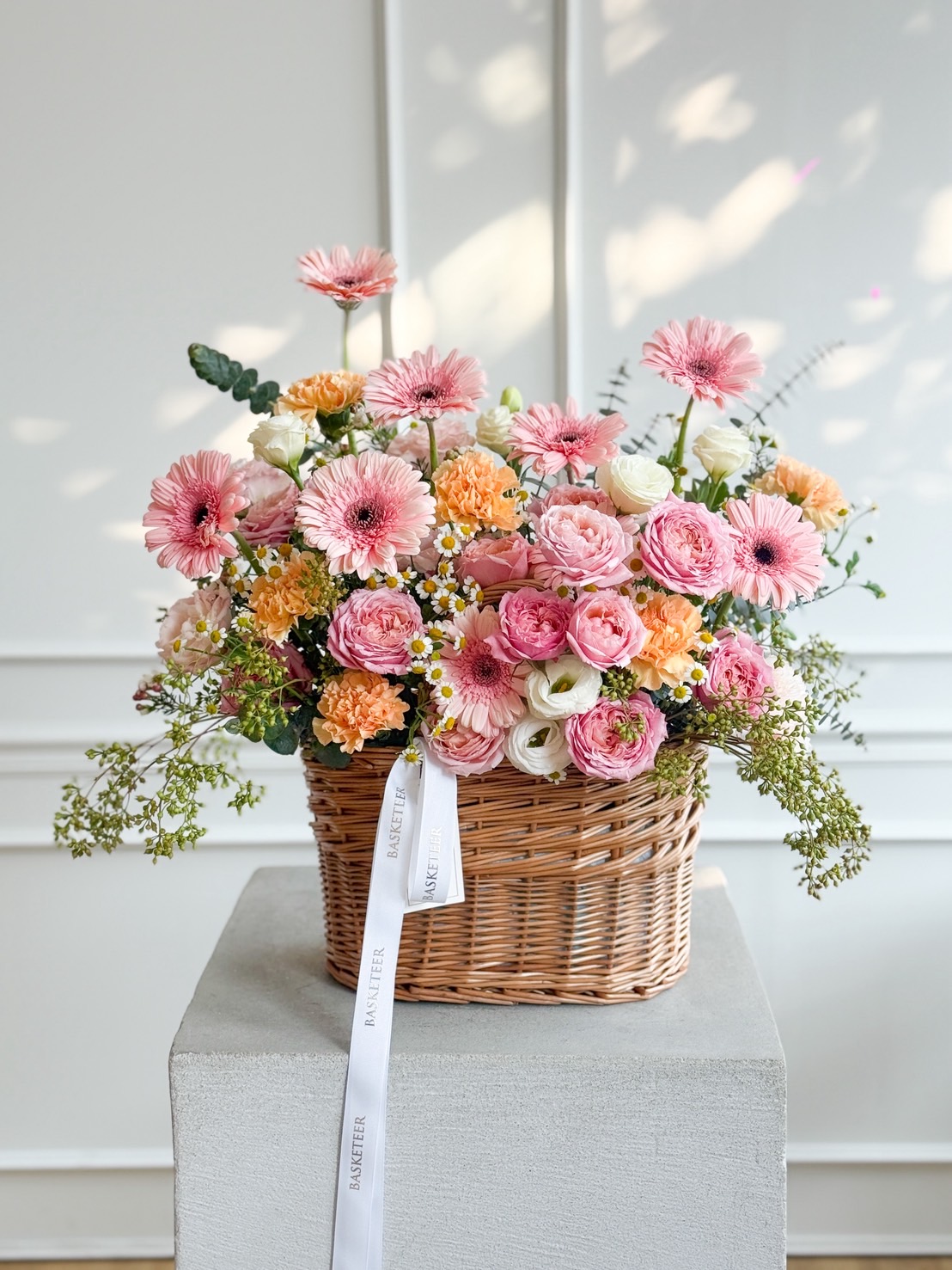 A wicker basket filled with a colourful arrangement of peach roses, white carnations, yellow gerbera daisies, small daisies, and greenery sits on a white pedestal against a light-coloured wall.
