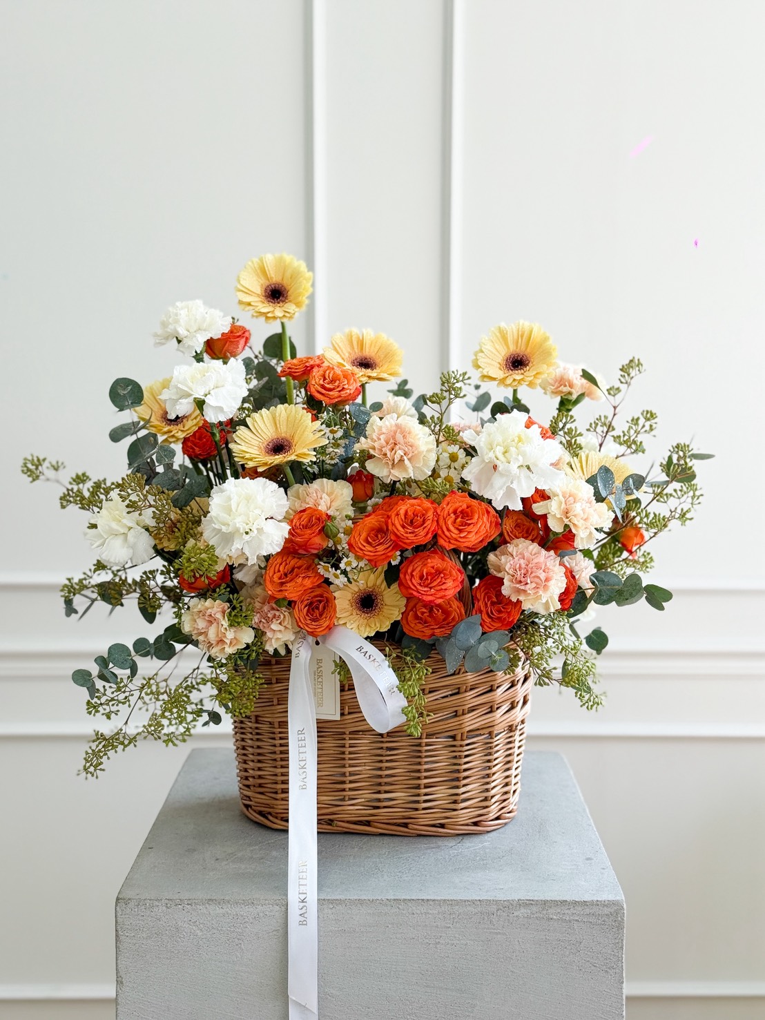 A wicker basket filled with Peach Roses, White Carnations & Yellow Gerbera Basket Bouquet sits on a grey pedestal against a white wall, adorned with lush greenery and finished with a white ribbon.