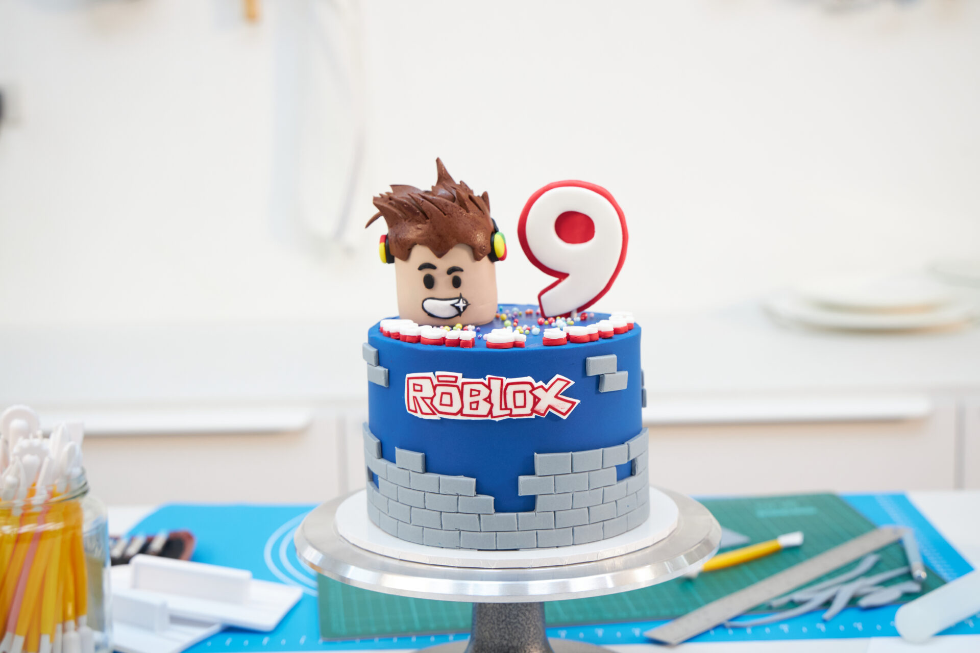 A classic blue Roblox birthday cake, topped with a character face and number nine candle, is ready for Charlotte’s celebration in Bangkok.