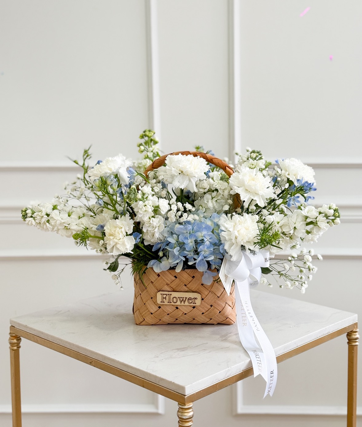 A woven basket filled with white carnations & blue hydrangea basket bouquet sits on a square marble table. A white ribbon adorns the handle, and 