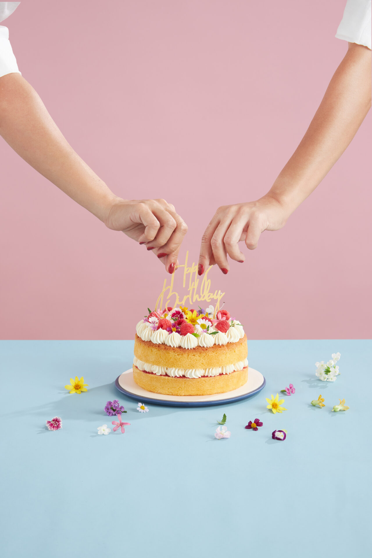 Two hands place a yellow “Happy Birthday” topper on a decorated cake with piped cream and flowers, on a blue surface scattered with small colourful flowers, against a pink background.