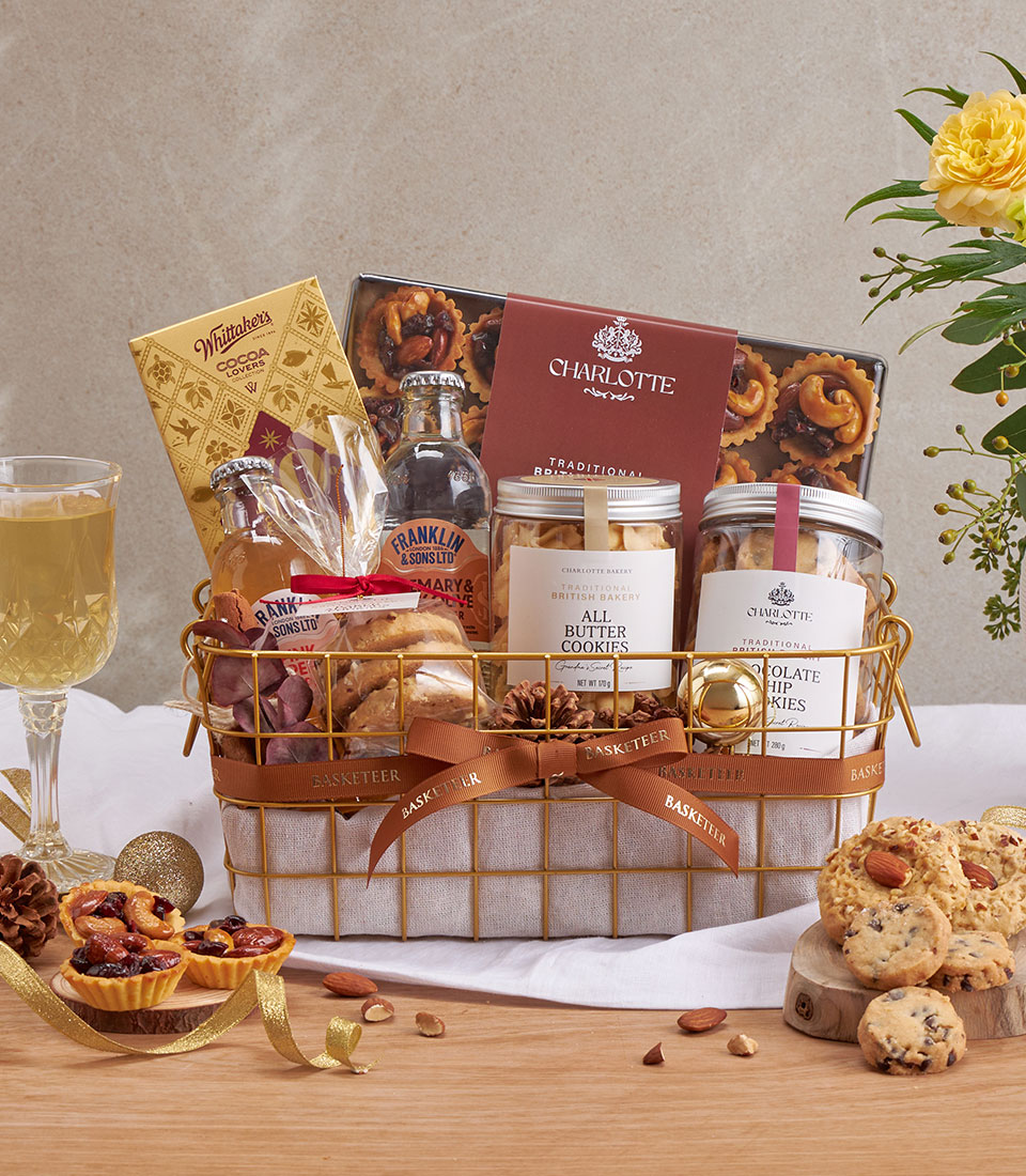 A gold wire gift hamper filled with gourmet treats, including biscuits, chocolates, pretzels, jam, a drinks bottle, and festive baked goods, displayed on a table with a glass of white wine and yellow flowers.