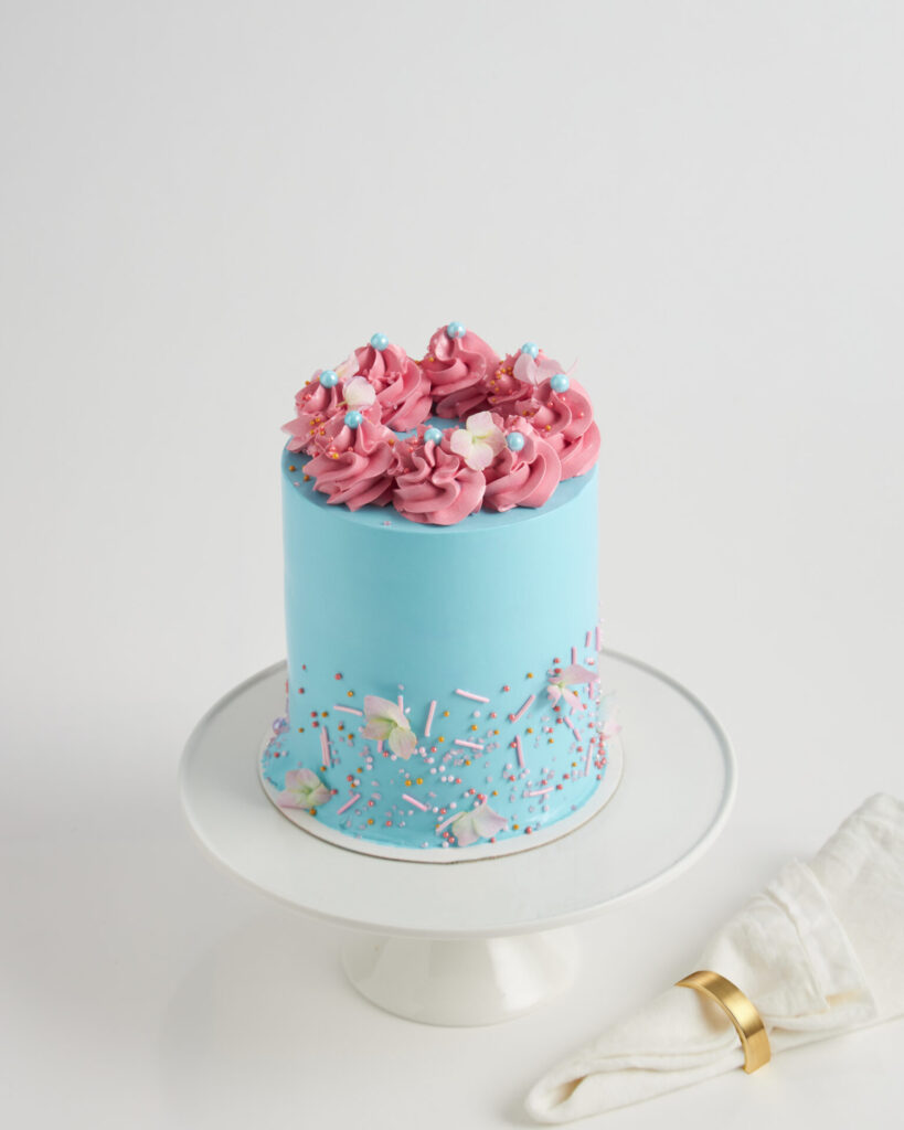 A light blue cake with pink icing swirls and sprinkles on a white stand, crafted to perfection by Charlotte Bakery Bangkok.