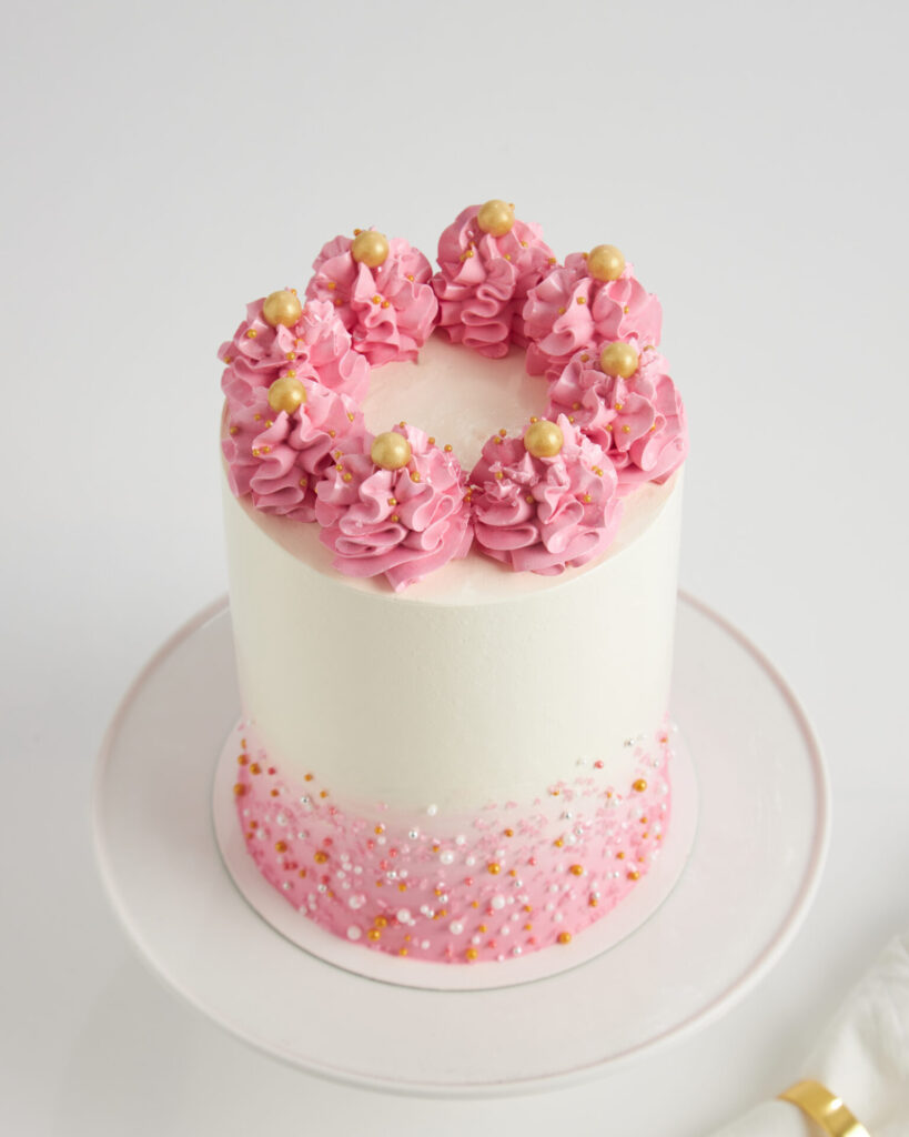 Charlotte Bakery Bangkok presents a tall white cake with pink icing swirls, gold pearls, and sprinkles on a classic white stand.