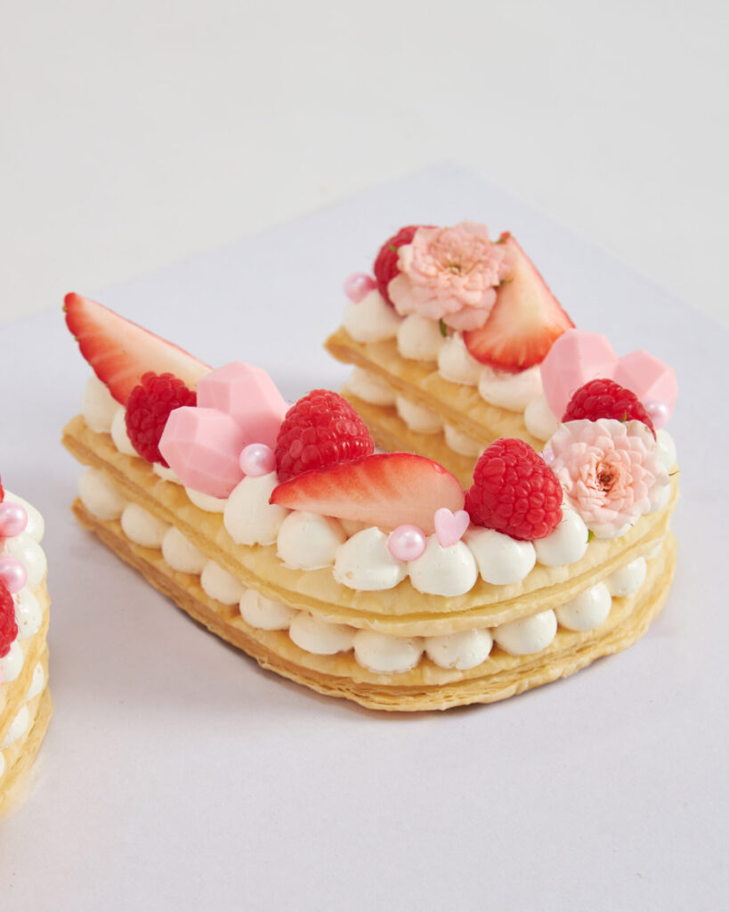 A heart-shaped pastry by Charlotte Bakery Bangkok, layered and topped with whipped cream, berries, pink sweets, pearls, and edible flowers.