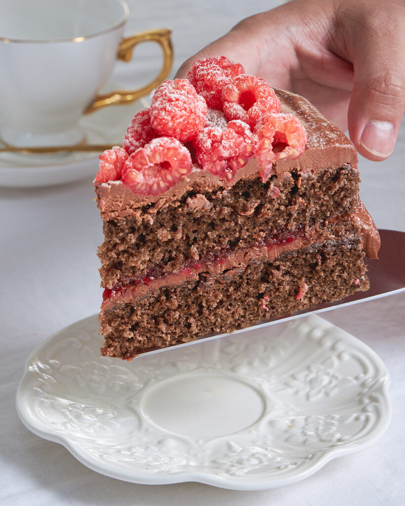 A slice of rich chocolate cake with glossy icing and raspberries is served on fine china beside a teacup at Charlotte Bakery Bangkok.