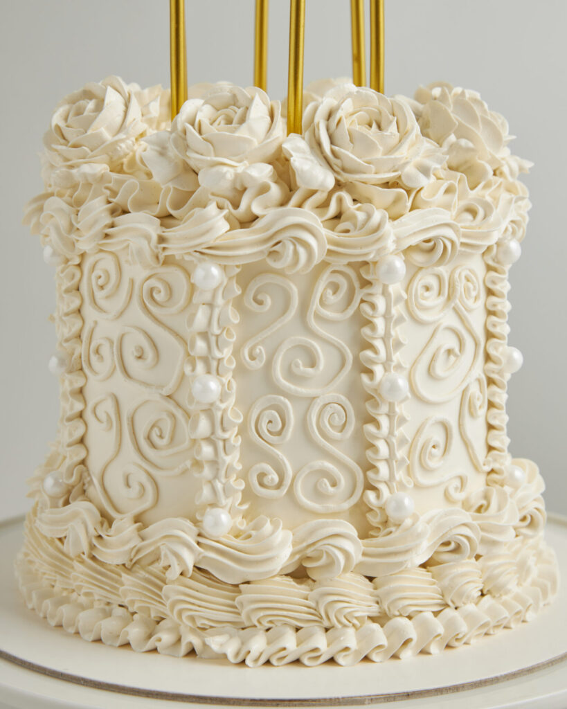 A tall, elegant white cake from Charlotte Bakery Bangkok features ornate swirls, rosettes, ruffled borders, pearls, and gold accents.