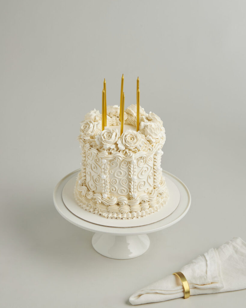 A white iced cake with delicate swirls and roses, topped by three gold candles, graces a white stand—Charlotte Bakery Bangkok.
