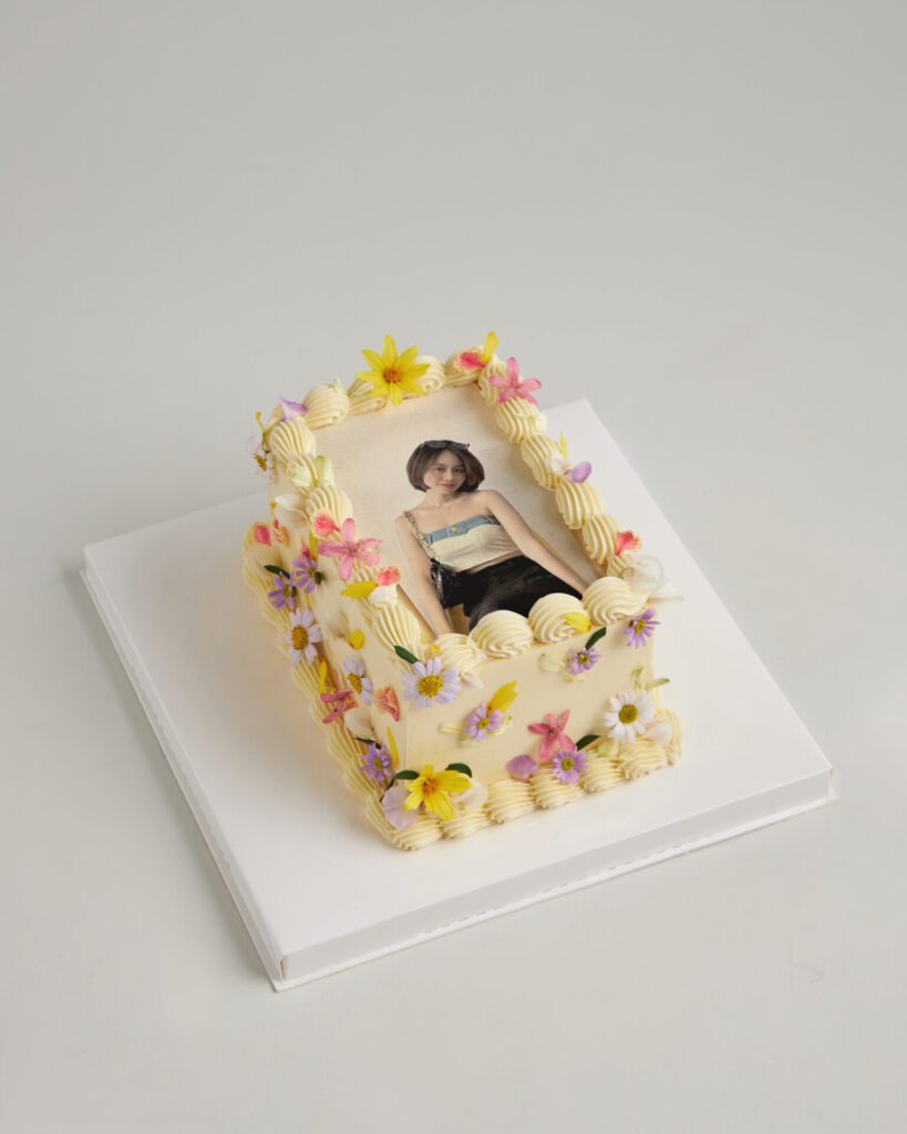 Rectangular Charlotte Bakery Bangkok cake with a woman's photo, piped white icing, edible flowers, on a white cake board.