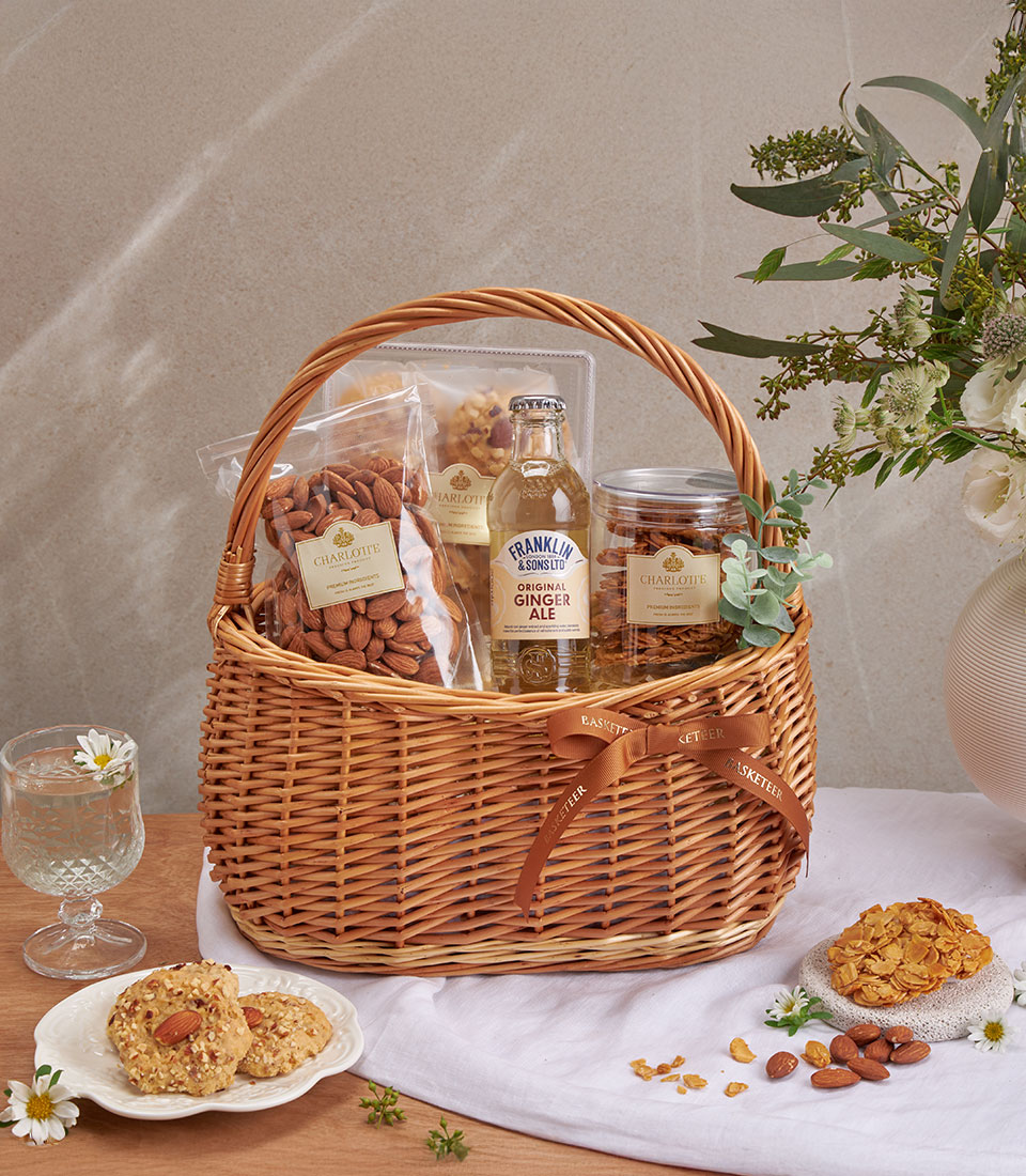 Full view of almond cookie & ginger ale basket with assorted snacks and drinks in a woven basket.