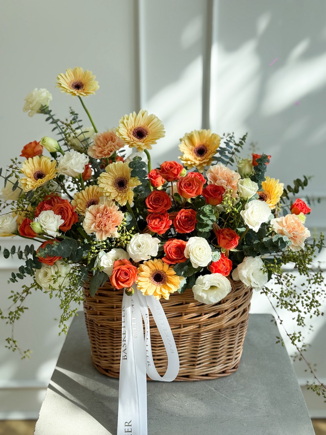 A wicker basket filled with a vibrant White Carnations & Yellow Gerbera Basket Bouquet and peach roses sits on a table, adorned with a white ribbon. Sunlight casts soft shadows over the cheerful arrangement.