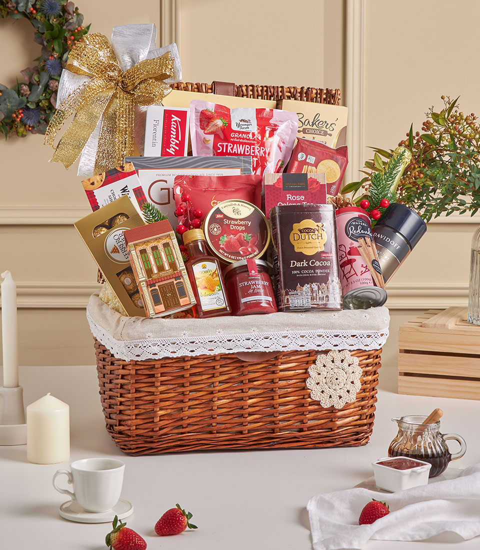 A Tea & Gourmet Snack Celebration Hamper, this wicker gift basket with lace trim and a gold bow is filled with deluxe chocolate, drinking chocolate, biscuits, jams, and assorted snacks. Displayed on a table with candles, strawberries, and a festive background.