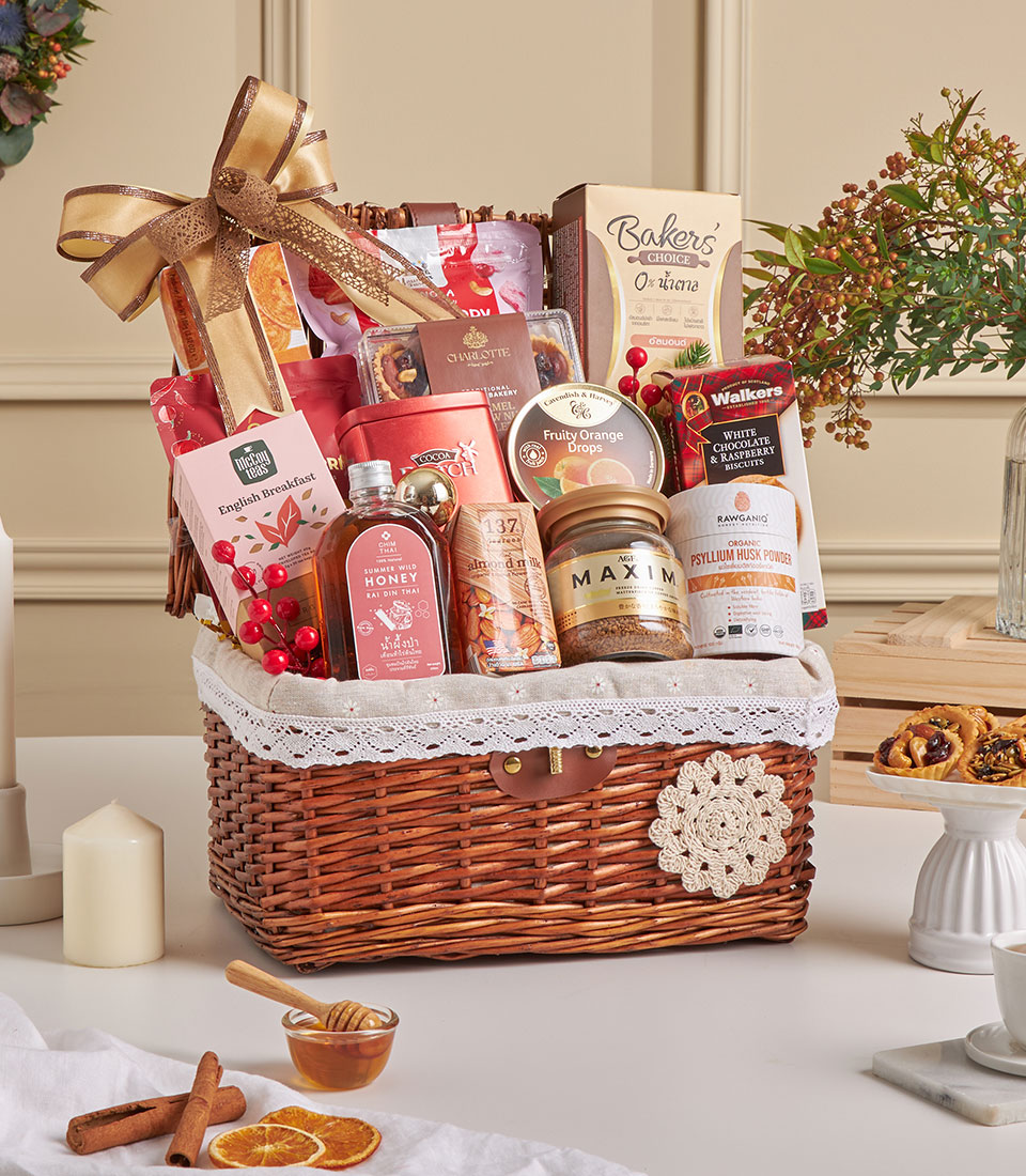 A wicker gift hamper filled with assorted delights like tea, honey, almond biscuits, chocolates, and snacks is decorated with a gold ribbon and festive berries. This Biscuits & Healthy Gourmet Treats Hamper sits on a white table beside a candle and dried orange slices.