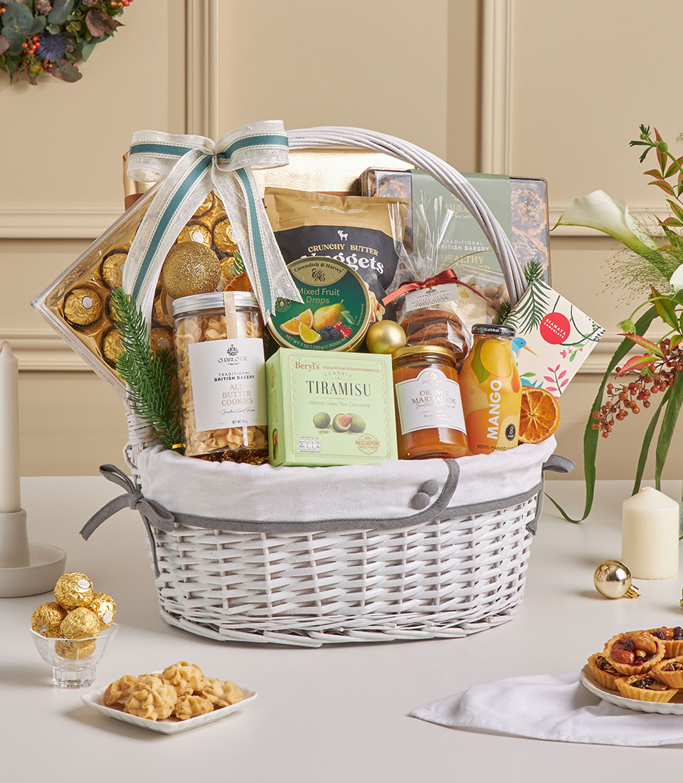 A white wicker Chocolate & Mixed Nut Gourmet Celebration Hamper, filled with chocolates, coconut roll, biscuits, tea, juice, tiramisu, and other delights, is adorned with ribbon and dried orange slices on a festive table with candles nearby.
