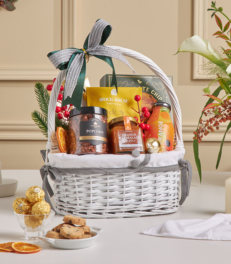 A white wicker hamper filled with snacks such as popcorn, biscuits, crisps, orange juice, jam, and decorated with a grey bow, faux berries, and greenery. In front are biscuits and gold-wrapped chocolates on a table.