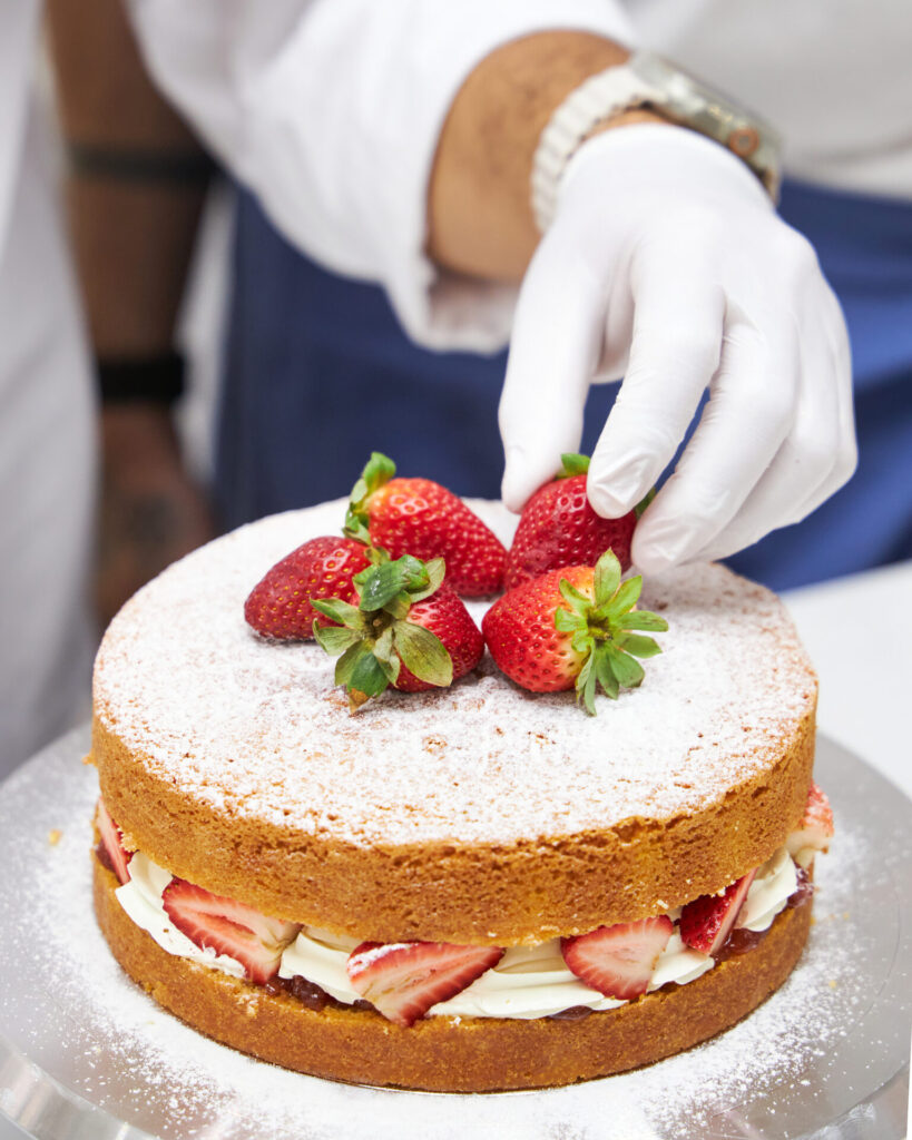 Chef ex, wearing white gloves, decorates a Victoria sponge cake by placing fresh strawberries on top of the powdered sugar–dusted cake
