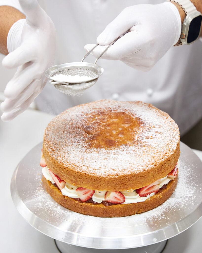 Chef dusting powdered sugar over a Victoria sponge cake layered with cream and fresh strawberries.