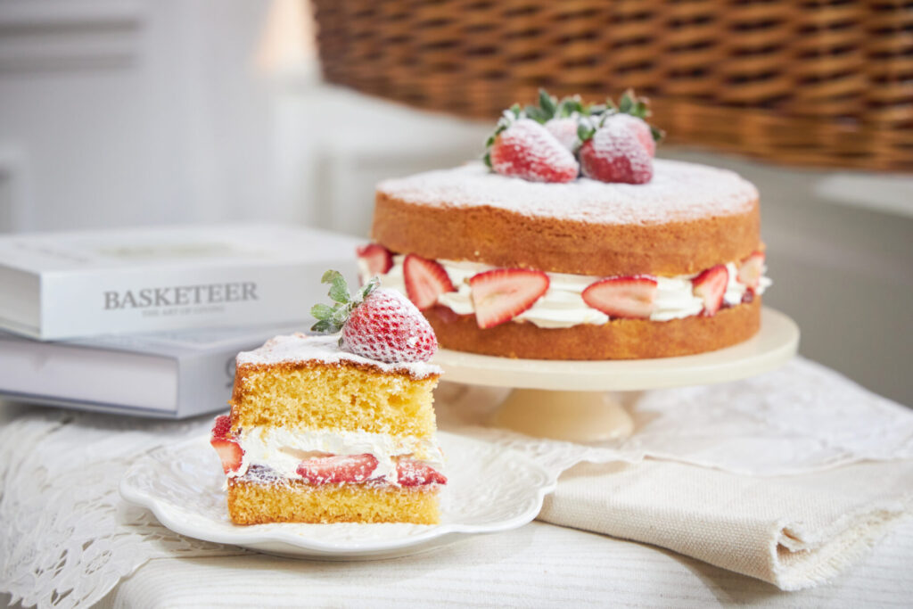 A slice of strawberry Victoria sponge cake placed on a white plate in front of a whole cake filled with fresh strawberries and cream, dusted with powdered sugar.