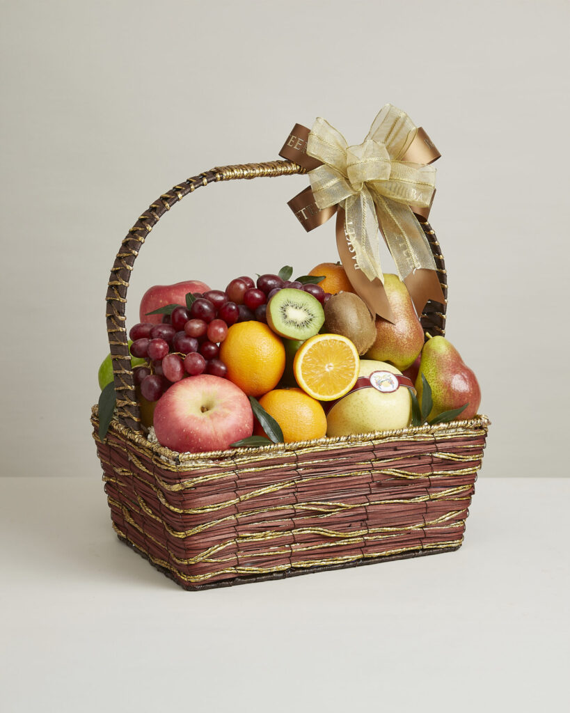 A wicker basket filled with assorted fresh fruit, including apples, oranges, kiwifruit, grapes, pears, and lemons, adorned with a large decorative gold and brown ribbon bow on the handle.