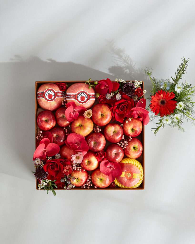 A square box filled with red and pink apples, some decorated with stickers, surrounded by red roses and other small flowers. A small vase with red and white flowers is placed to the right of the box.
