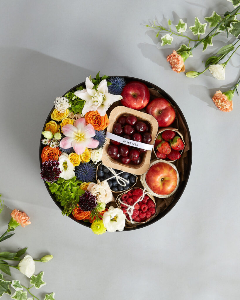 A premium round gift tray filled with fresh fruits and vibrant flowers from Basketeer, featuring cherries, apples, berries, and mixed blooms.