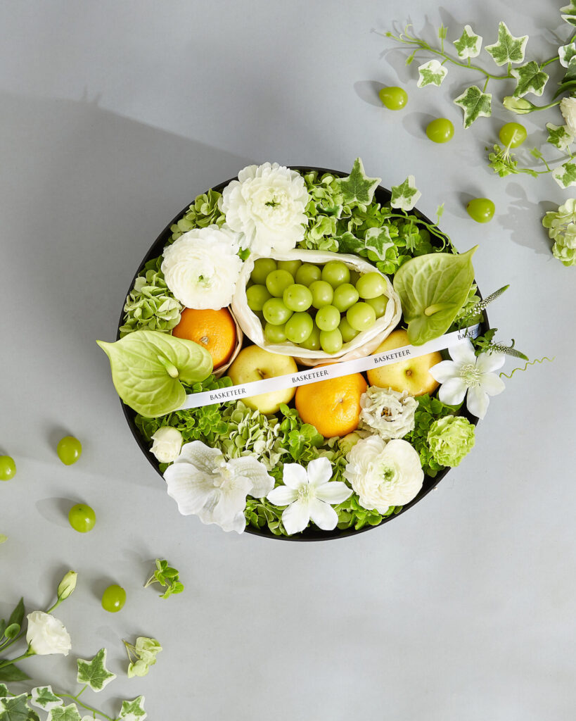 A premium green and white fruit and flower gift tray from Basketeer featuring Shine Muscat grapes, apples, oranges, and elegant white florals.