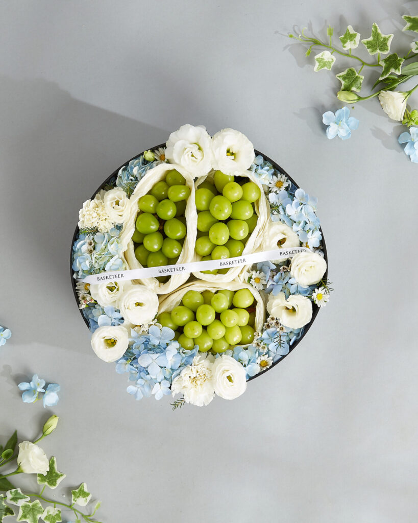 Luxury round gift box filled with Japanese Shine Muscat grapes surrounded by pastel blue and white premium flowers.
