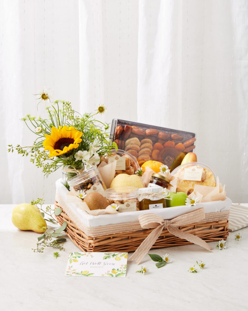A wicker gift hamper with a bow, filled with fresh fruit, snacks, jars, and a bouquet with a sunflower, sits on a white surface next to a "Get Well Soon" card and a few flowers.