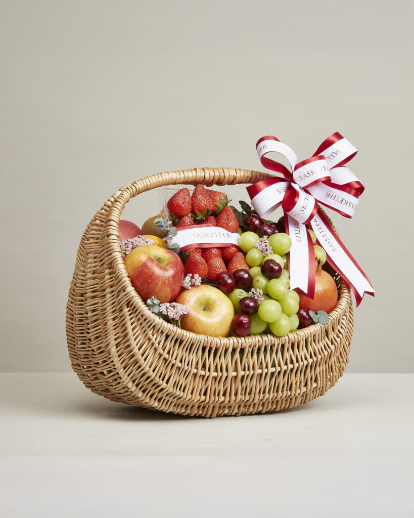 Premium congratulations fruit basket filled with strawberries, apples, green grapes, and seasonal fruits wrapped with a red and white ribbon from Basketeer.