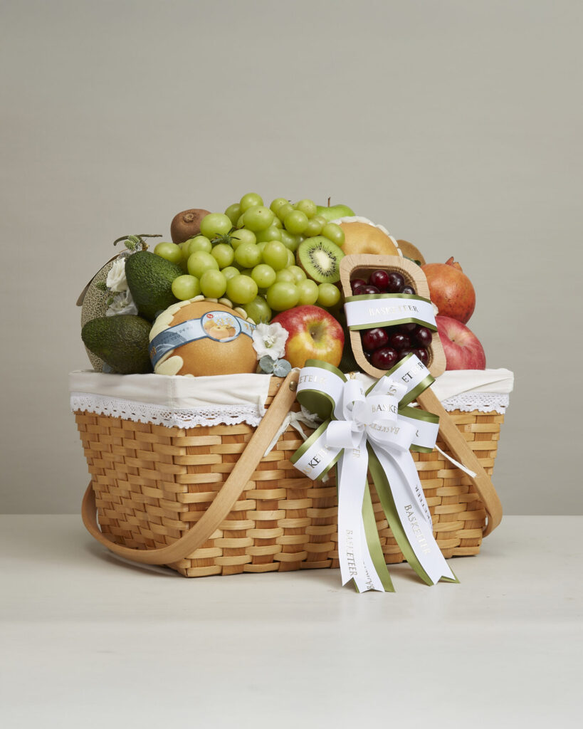 A wicker basket filled with assorted fresh fruit, including grapes, kiwifruit, pomegranates, avocados, melons, and cherries, decorated with a white lace cloth and a bow with white and green ribbons.