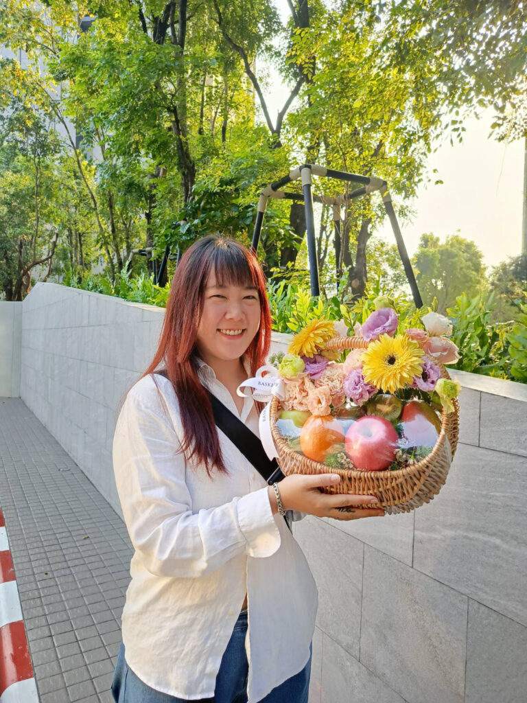 A smiling woman with long hair, wearing a white shirt, stands outdoors holding a wicker basket filled with bright flowers and apples. Green trees and a stone wall line the sunny background.