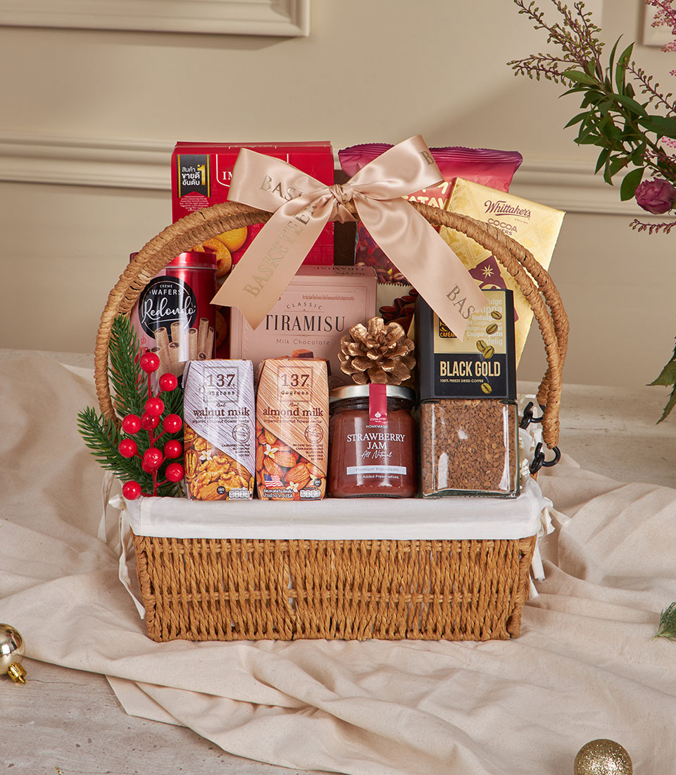 A champagne gold wicker holiday hamper filled with tiramisu, cookies, coffee, and assorted festive treats arranged with a soft gold ribbon, red berries, and holiday décor