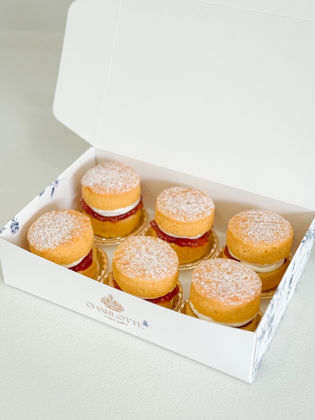 An open white box Six round pastries filled with cream and jam, topped with icing sugar, arranged in two rows inside an open white bakery box.