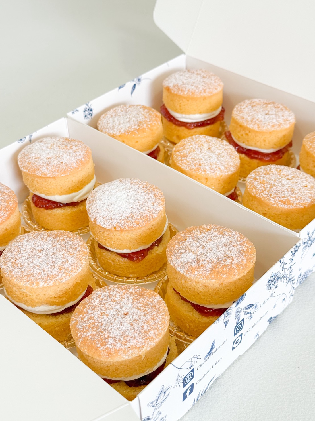 A box of twelve mini sponge cakes, each filled with cream and strawberry jam, and topped with a dusting of icing sugar. The cakes are neatly arranged in two rows inside a white patterned box.